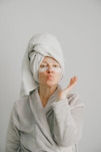 Woman with a towel on her head and eye masks, blowing a kiss in a robe.