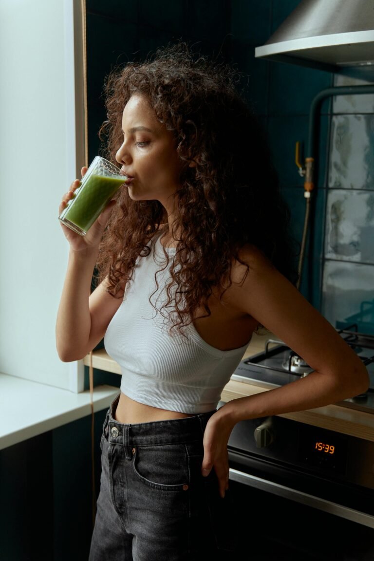 Young woman with curly hair drinks green smoothie indoors. Emphasizes healthy lifestyle.