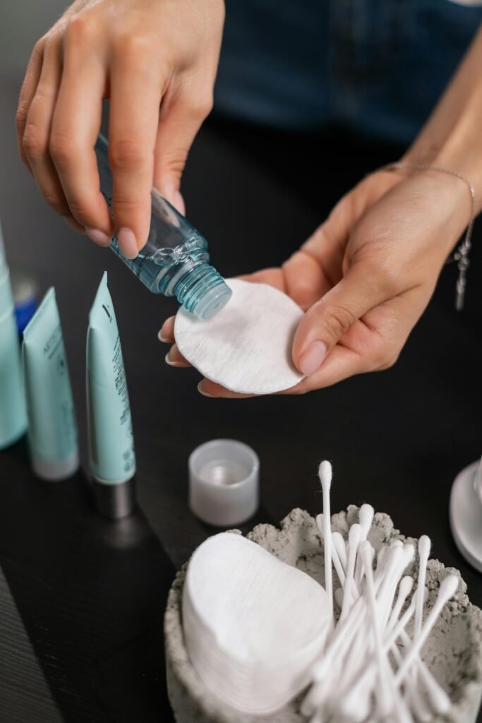 Close-up of hands applying toner to a cotton pad with skincare products in the background.